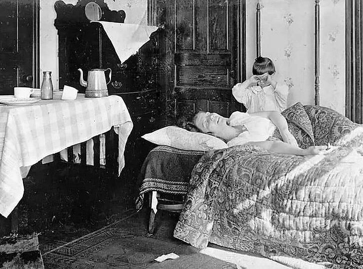 १९१८ नोभेम्बर को तस्विर । स्पेनिस फ्लूकी बिरामी एक महिला बेडमा सुतिरहेकी छिन् । साथमा उनकी छोरी छटपटाइहरेकी छिन् ।